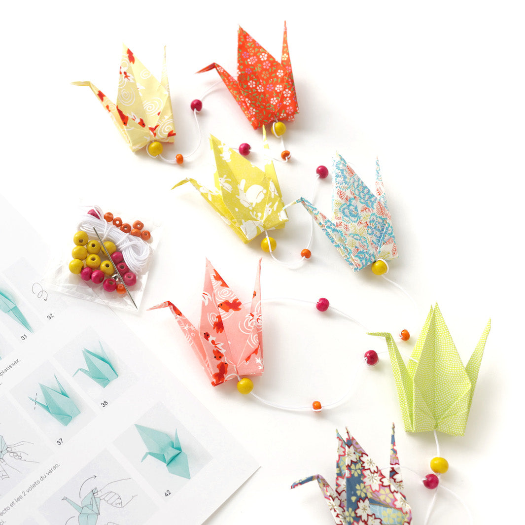 vue zoomé de la guirlande montée et du dépliant du kit créatif guirlande de grues en origami « granité » (K9) dans les tons jaunes, rouge orangé, roses, vert acidulé, bleus et mauve